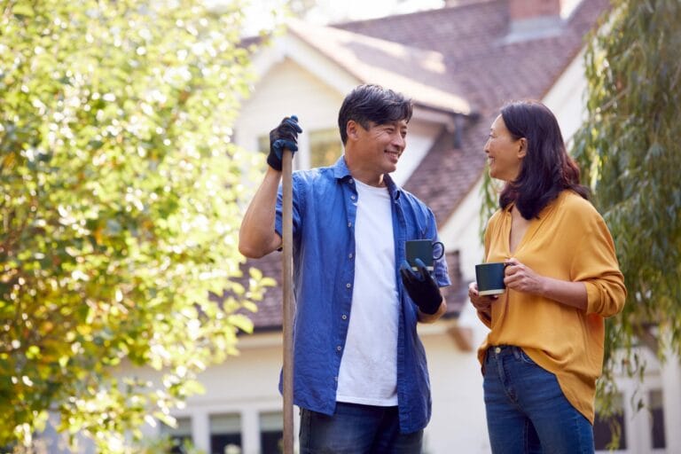 Photo of a man and a woman smiling outside with mugs in their hands. The man is holding a shovel.