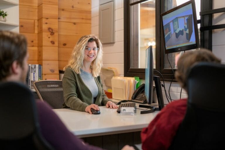 Photo of a woman smiling and showing two people a screen while on her computer.
